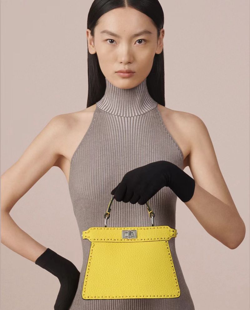 Woman poses indoors with a bright yellow leather Fendi Peekaboo bag, wearing a grey turtleneck dress and black gloves against a neutral backdrop.