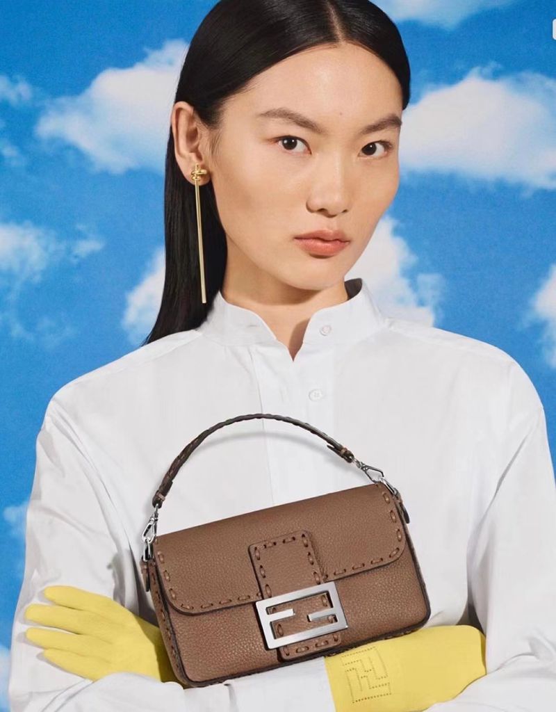 Portrait of a woman in a white shirt and yellow gloves holding a brown leather handbag with a silver logo against a sky backdrop.