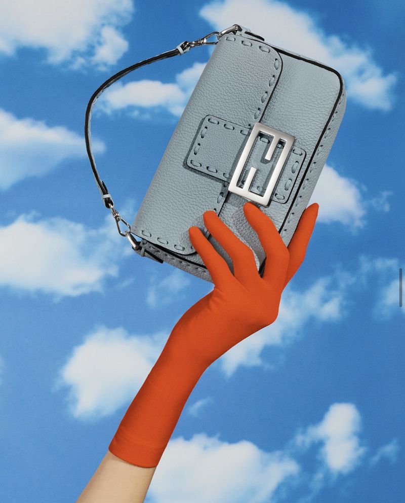 Fashion shot of a light blue Fendi handbag with a silver logo clasp, held by a hand wearing an orange opera glove against a cloudy sky.