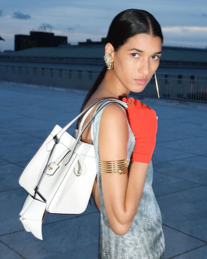 Fashionable woman wearing an elegant dress and striking jewelry, carrying a modern white tote bag, photographed outdoors against a city backdrop.