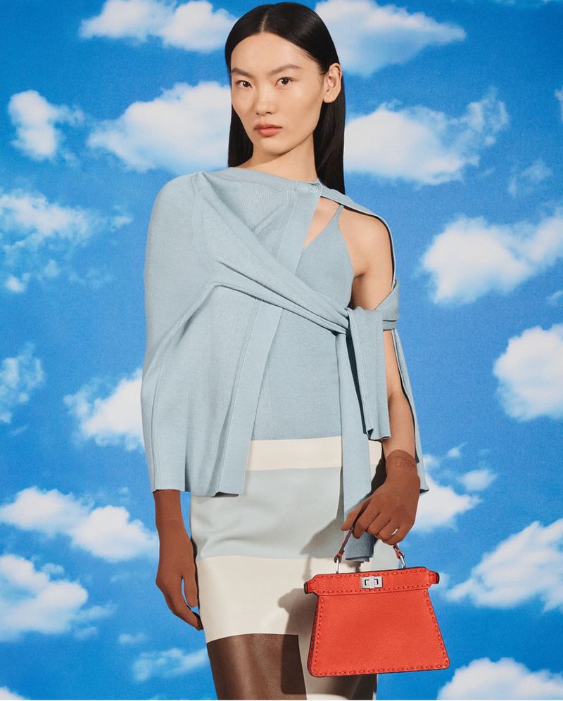 Asian woman wearing a blue top and light skirt with an orange handbag poses against a light blue sky background in a fashion shot.