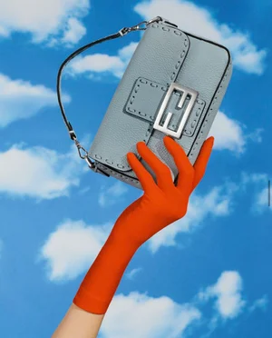 Hand in a long orange glove holding a light blue Fendi handbag against a bright sky with white clouds.