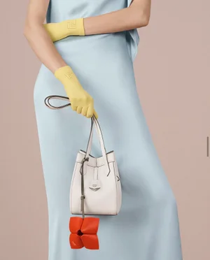 Model in pale blue dress wearing yellow gloves holding white handbag with orange charm against plain background