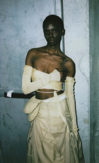 Fashionable African American woman in long beige dress and matching long fingerless gloves poses indoors against a plain wall.