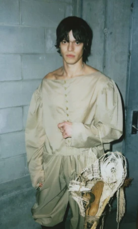Portrait of a young adult in a historical-style beige-toned outfit holding a macrame-like object in front of a concrete-block wall.