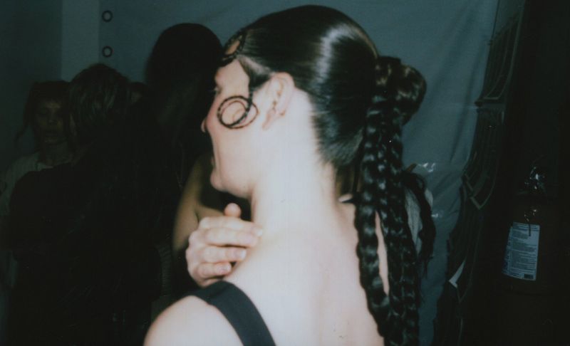 Woman with braided ponytail and styled dark hair indoors, partial view of people and an extinguisher in the blurred background.
