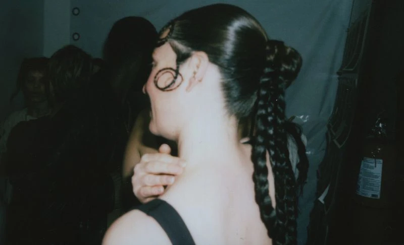 Woman with braided ponytail and styled dark hair indoors, partial view of people and an extinguisher in the blurred background.