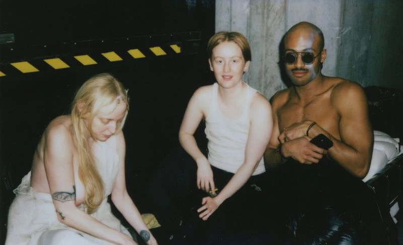 Three people pose in a dimly lit, indoor environment. Two are seated, one standing, with dark walls and a yellow striped safety barrier.