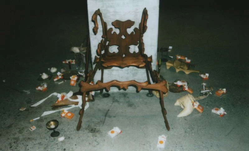 Ornate wood chair stands amidst scattered paper debris on a gray floor against a neutral wall in this mysterious indoor scene.
