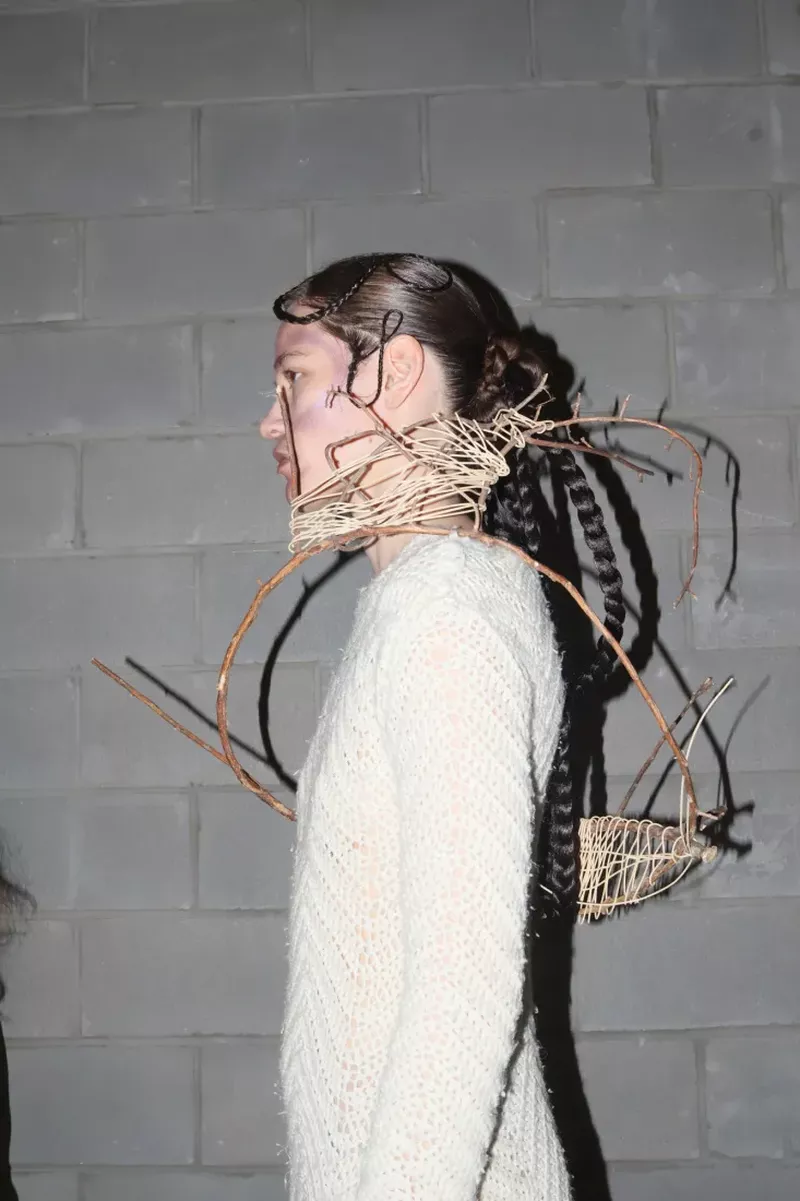 Adult female fashion model in profile wearing a white sweater with woven headpiece and dark braided hair against a concrete brick wall.