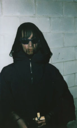 A person with black sunglasses, hooded cloak, and visible 'blood' in front of a white brick wall, holding a small cross.