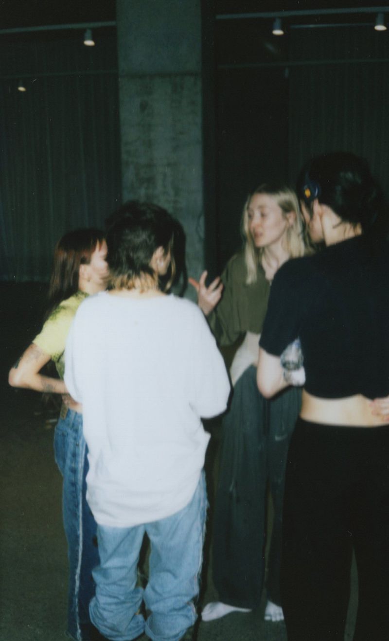 A group of four women stand together in casual clothing, possibly in a studio or industrial setting with neutral colors and diffused lighting.