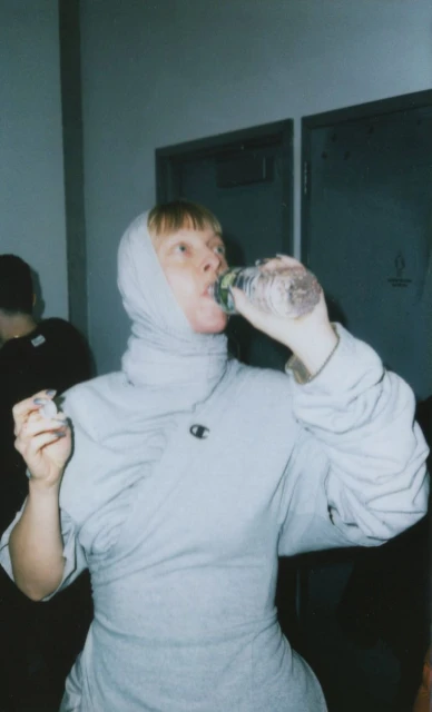 A woman in a light gray outfit and head covering drinks from a clear plastic water bottle indoors with a neutral background and closed doors.