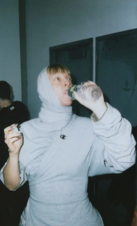 A woman in a light gray outfit and head covering drinks from a clear plastic water bottle indoors with a neutral background and closed doors.
