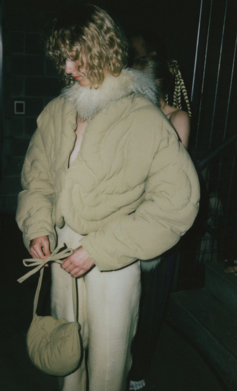Young adult poses indoors wearing a tan puffer jacket with fur collar, tied pants, and a matching bag, with a woman in the background.