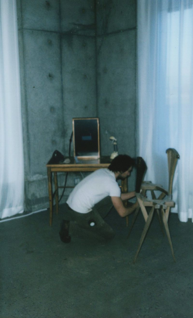 An adult male is crouched over a light wood chair, possibly repairing it, with a table, mirror, and curtain in the background indoors.