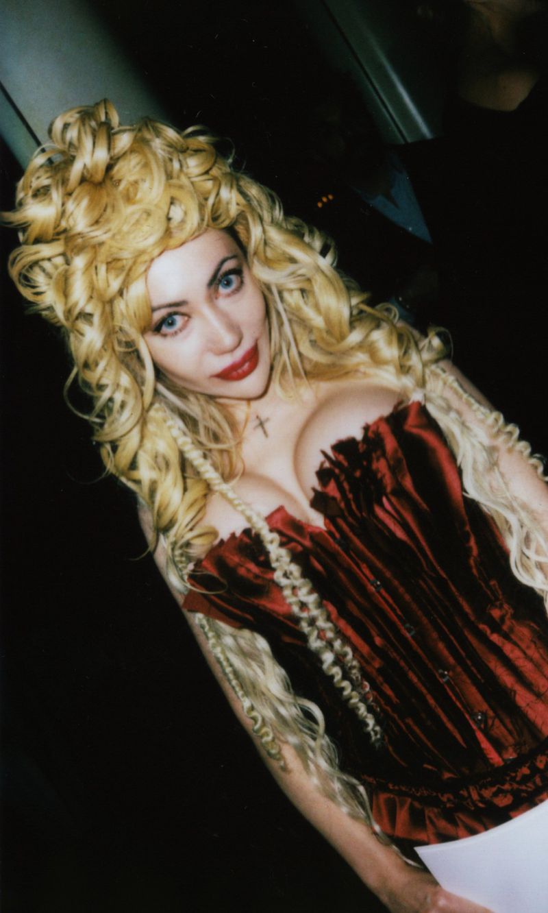 Portrait of a blonde woman wearing a red corset and a cross necklace against a dark background. 