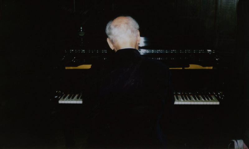 Back view of an elderly adult male playing a large dark wood-paneled electric organ with visible keyboards, set in a dimly lit room.