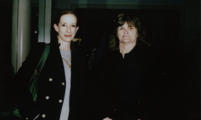 Two adult women pose indoors with dark clothing and a green shoulder bag, creating a candid and slightly grainy vintage-style portrait.