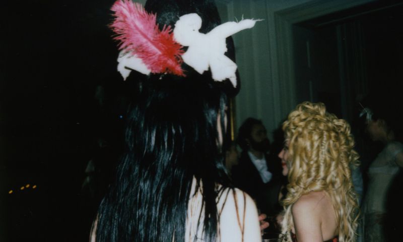 Two adult women at an indoor event with dark hair, blonde curly hair, feather hair accessories, and a blurred group in the background.