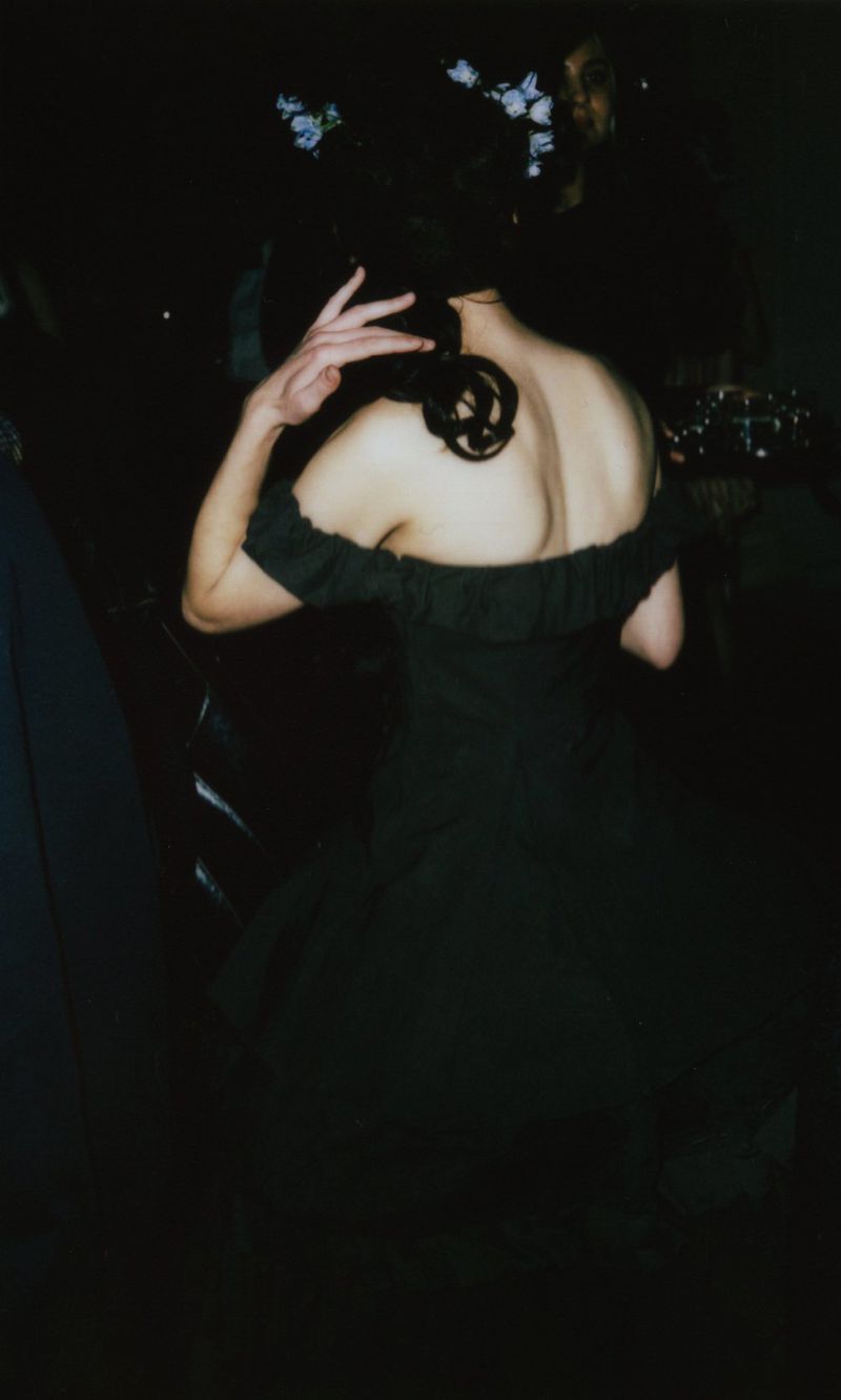 Woman in off-the-shoulder dress with floral hairpiece, standing indoors in low light with hand raised toward her hair, back view.