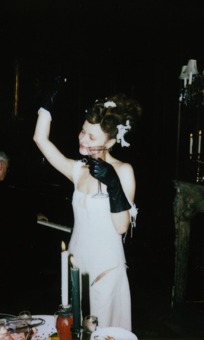 Woman in a white dress and black gloves raises a glass in a dimly lit room with a table, candles, and architectural details.