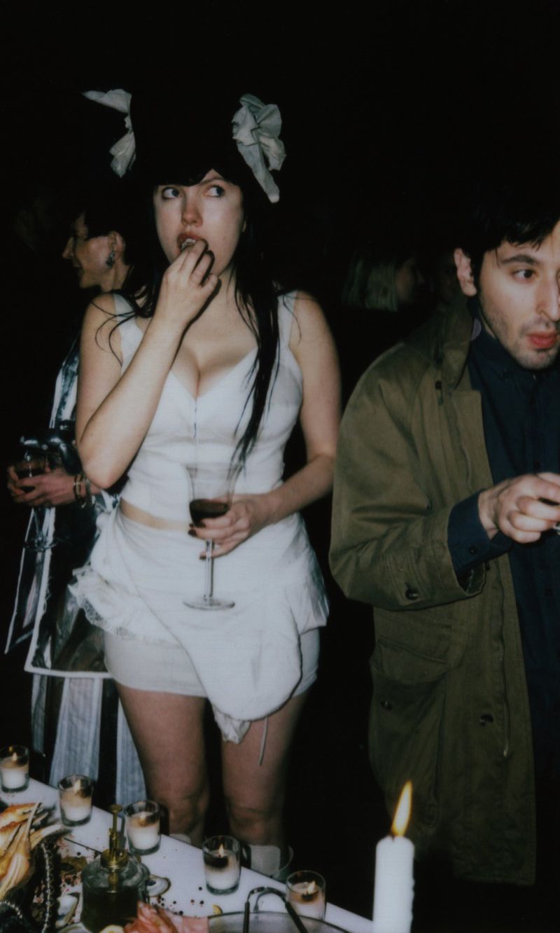 Woman in costume holds wineglass near table with snacks and candlelight with blurred people in background in a dimly lit setting.
