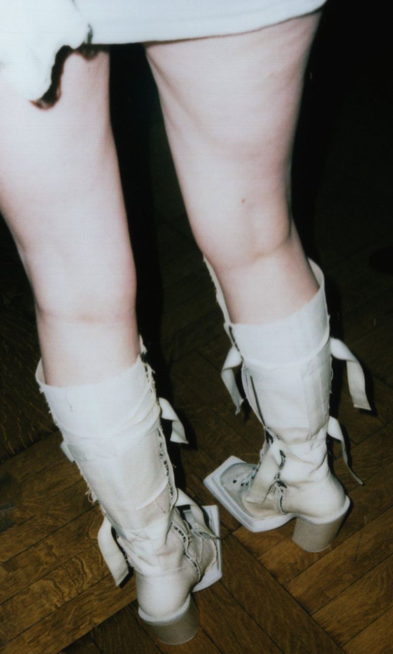 Close-up of a human from the knees down, wearing light-beige platform boots with a lace-up front, ribbon details, and high heels on a wood floor.