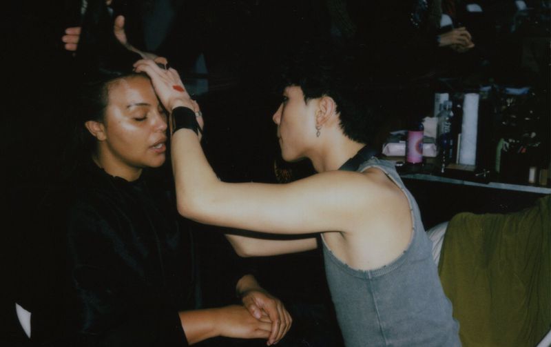Makeup artist applies makeup to the face of a female model in a dimly lit setting; beauty and fashion shoot.