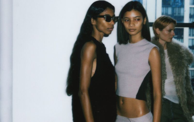 Three fashion models pose in front of a window with city buildings in the background, wearing crop tops and jackets.