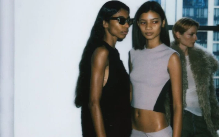 Three fashion models pose in front of a window with city buildings in the background, wearing crop tops and jackets.