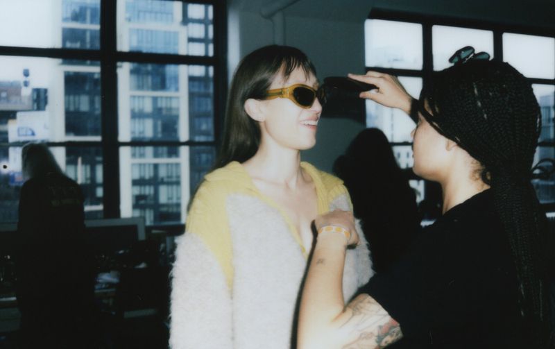 Woman wearing sunglasses smiling in an indoor setting while another woman adjusts her hair with an urban background visible through a window.