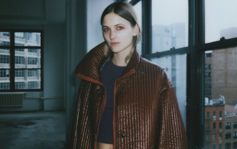 Adult woman posing in a high-rise apartment wearing a brown quilted jacket. Natural light with muted color palette and urban background.