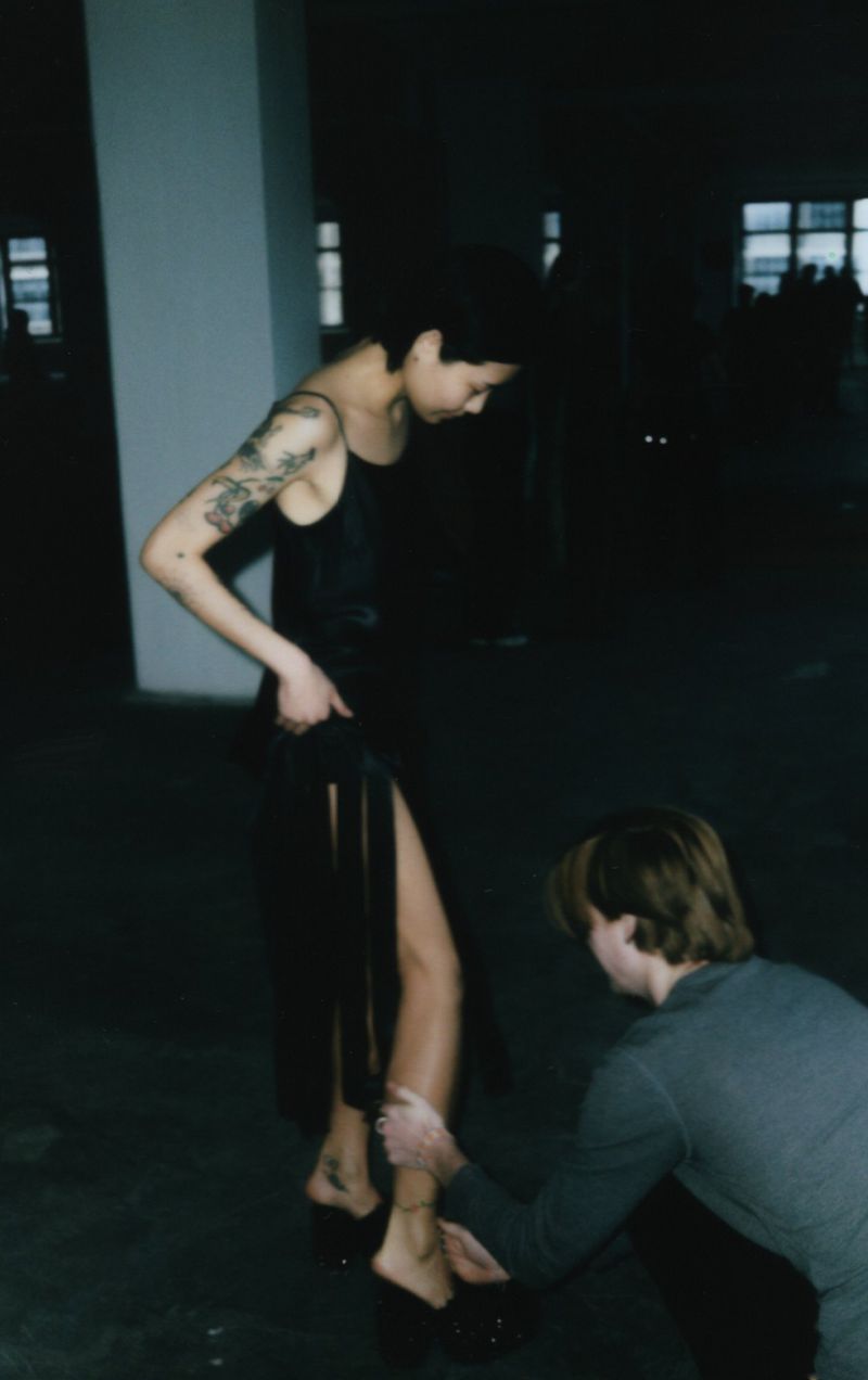 An adult female model in a dress is being helped by an adult male in a dimly lit indoor environment. The model has tattoos on her arm.