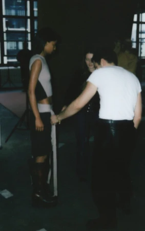 Fashion shoot with a model being measured by stylists in a studio, featuring an adult female wearing a crop top, mini skirt and platform boots.