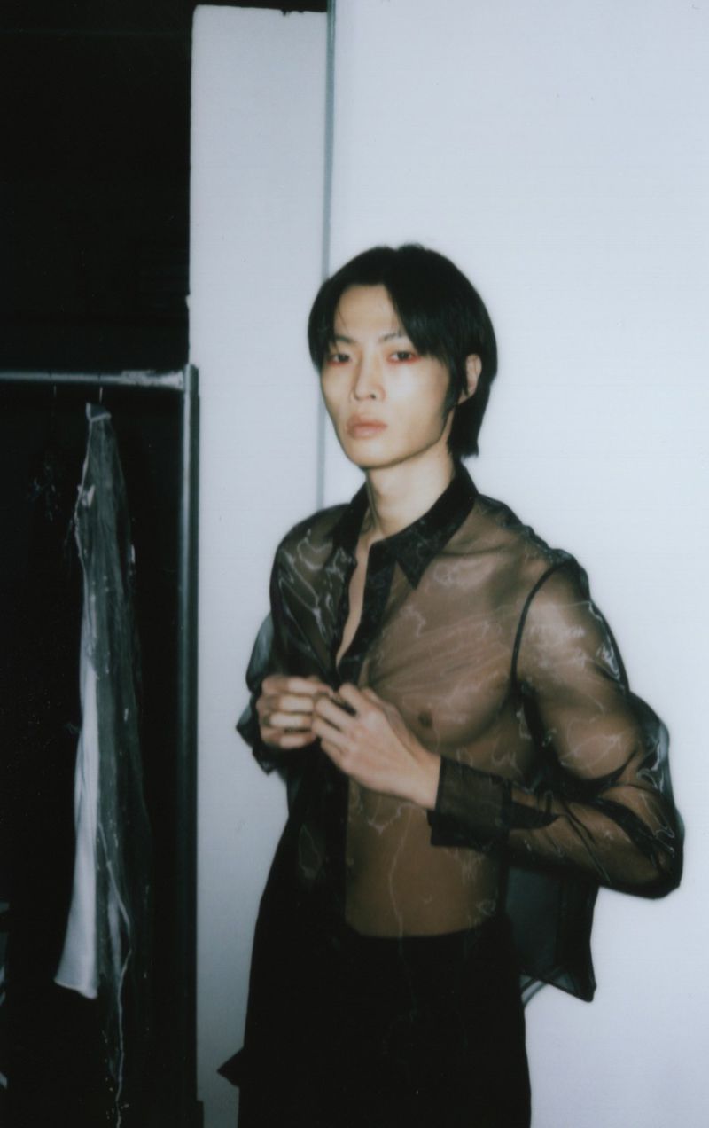 Portrait of an adult man in a sheer top adjusting his buttons in a dimly lit room, with black pants and medium dark hair.