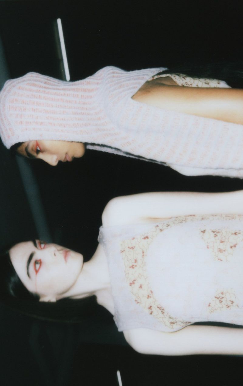 Two fair-skinned fashion models pose indoors. One wears a knitted balaclava, the other a frilly dress with floral details against a dark backdrop.