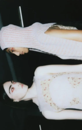 Two fair-skinned fashion models pose indoors. One wears a knitted balaclava, the other a frilly dress with floral details against a dark backdrop.