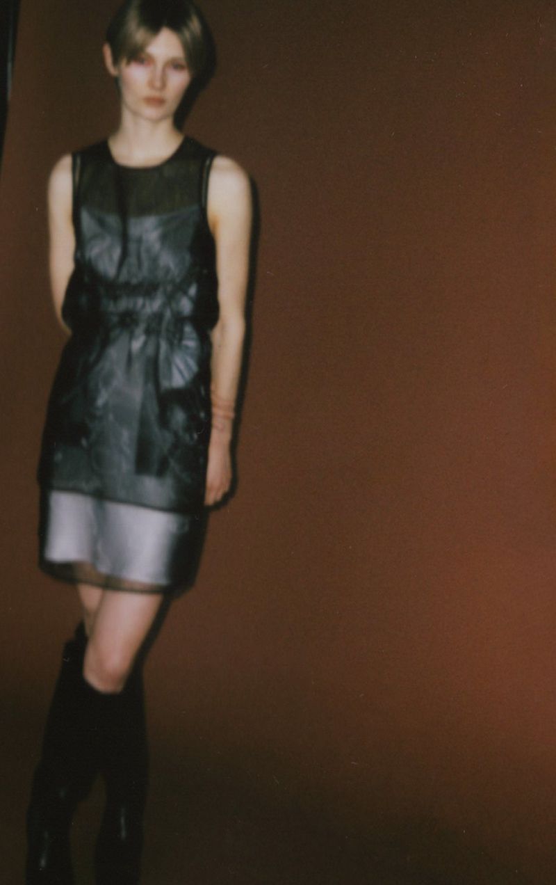 A woman poses in a sleeveless black dress with knee-high boots against a brown studio backdrop, presenting a high-fashion editorial aesthetic.