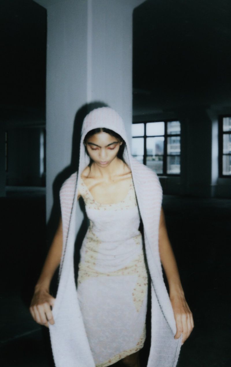 Woman poses in a sleeveless embellished dress and hooded wrap, standing in a dimly lit industrial space with large windows and concrete columns.