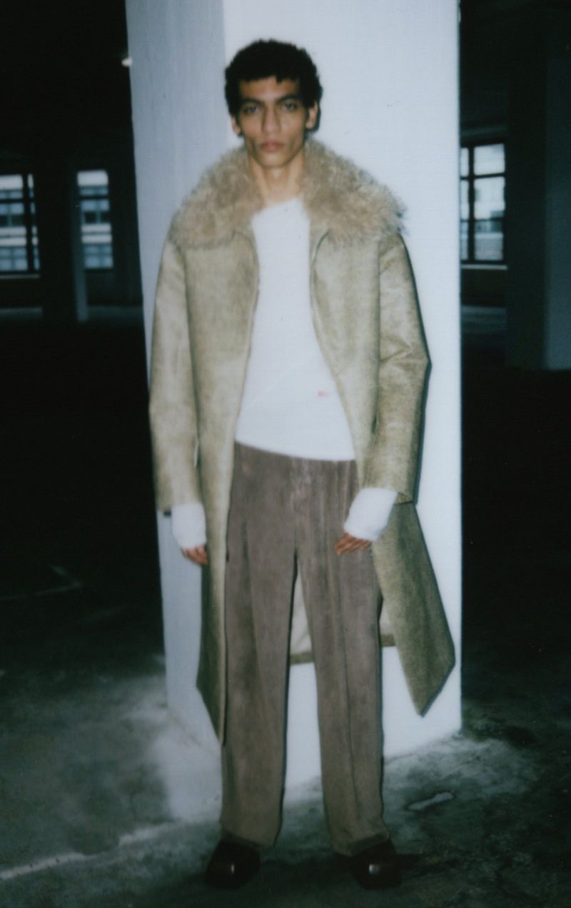 A model poses in a light beige shearling coat, white sweater, and brown pants indoors with white walls, brown flooring, and natural lighting.