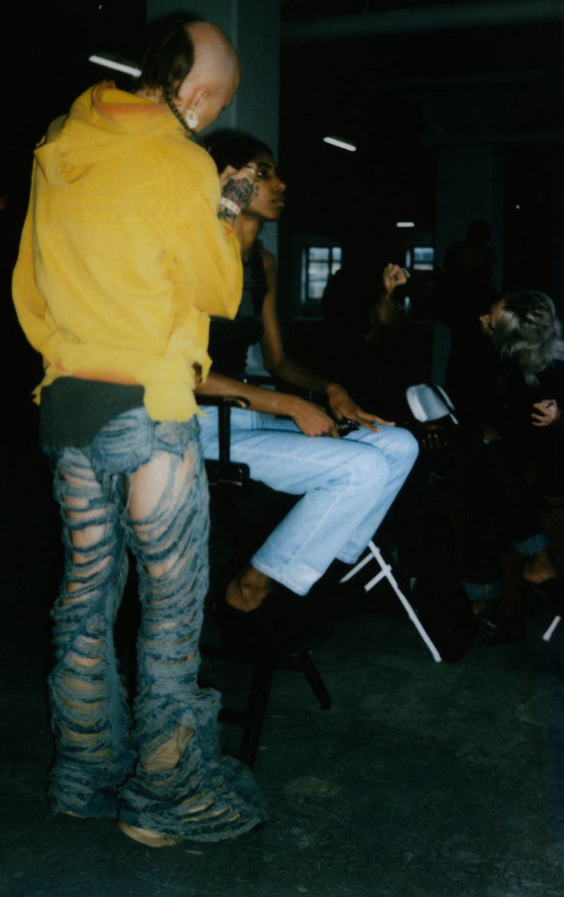 Two people in an indoor setting, one wearing a yellow hoodie and shredded jeans, the other seated in jeans, under dim lighting.
