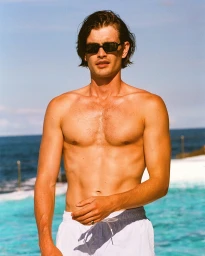 Shirtless man wearing dark sunglasses and white swim trunks standing outdoors near a blue ocean backdrop