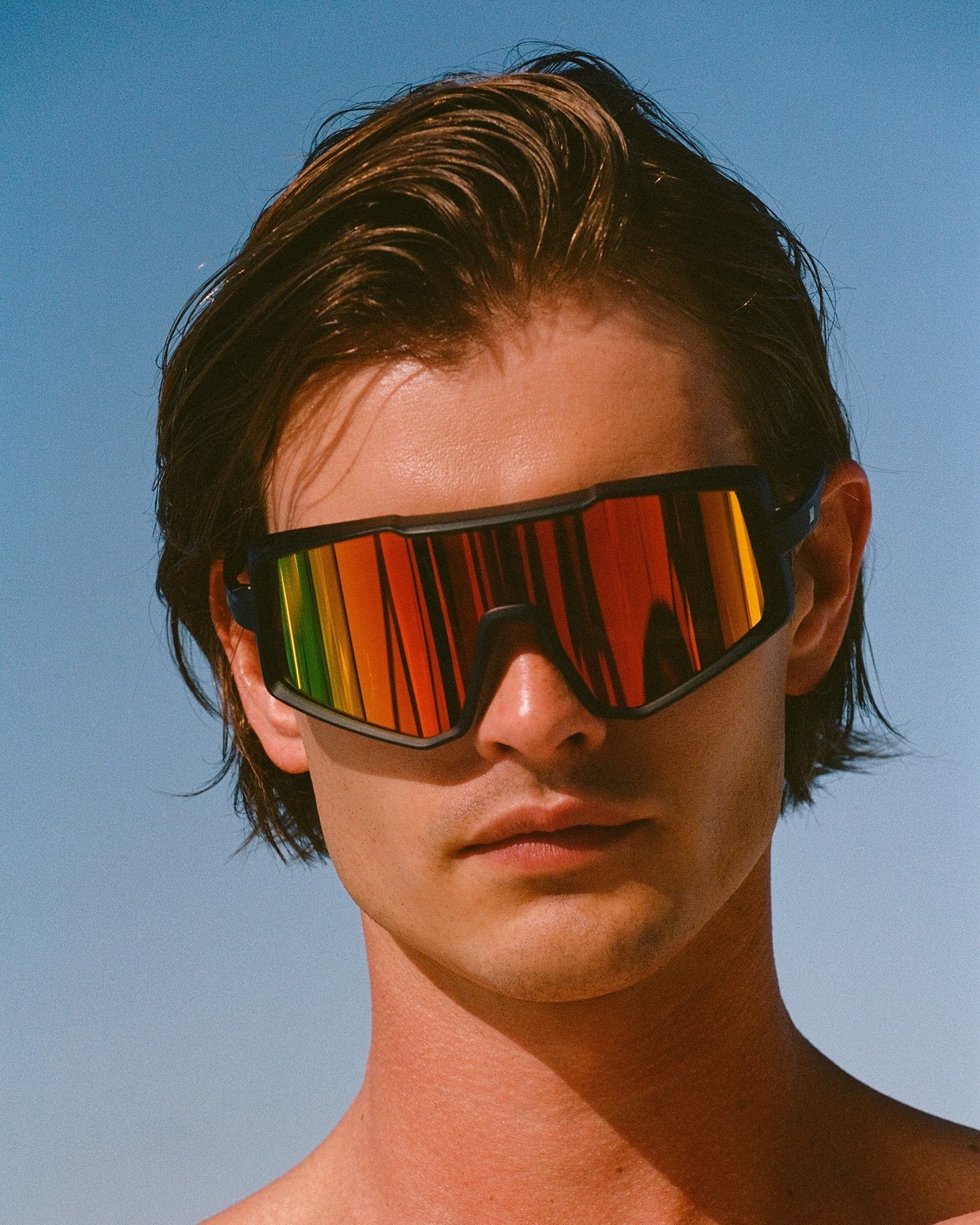 Close-up portrait of a young adult male wearing futuristic-style black sunglasses with rainbow mirrored lenses against a light blue sky.