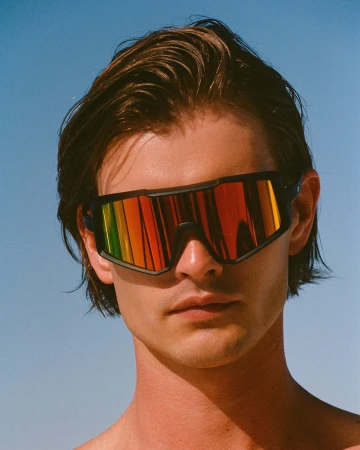 Close-up portrait of a young adult male wearing futuristic-style black sunglasses with rainbow mirrored lenses against a light blue sky.