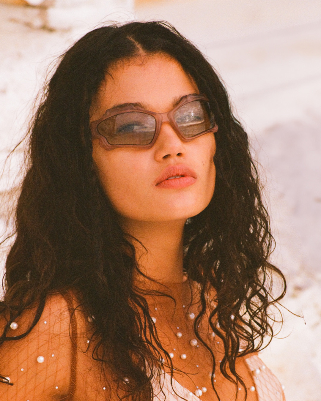 Woman with dark curly hair wears statement sunglasses, adorned pearl necklace in soft focus on a white architectural background.