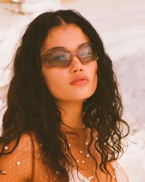 Close-up portrait of a woman with dark wavy hair wearing translucent sunglasses and pearl-embellished top outdoors.