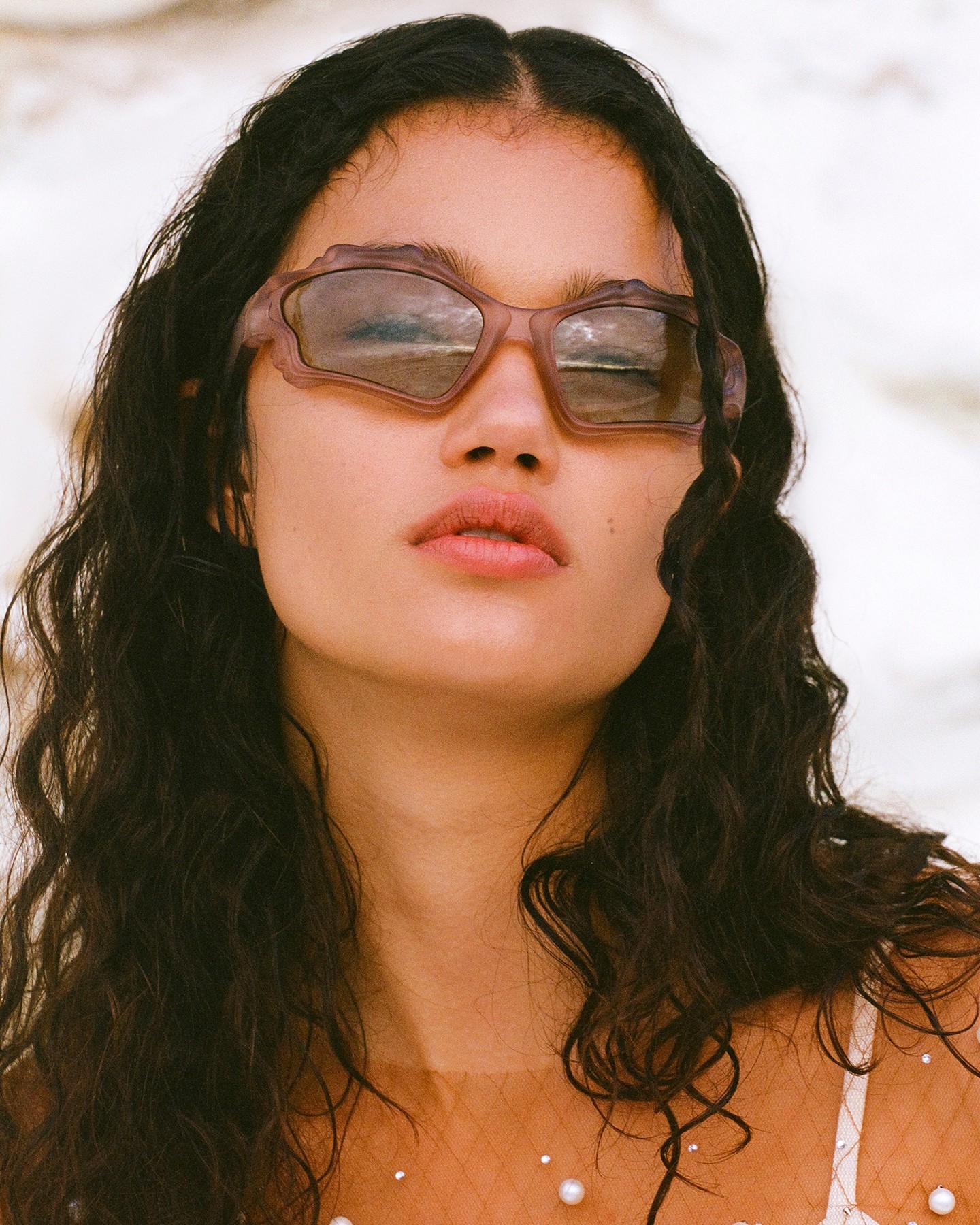Close-up of a young woman wearing mauve sculptural sunglasses, pearls, and a translucent top against a textured white background.