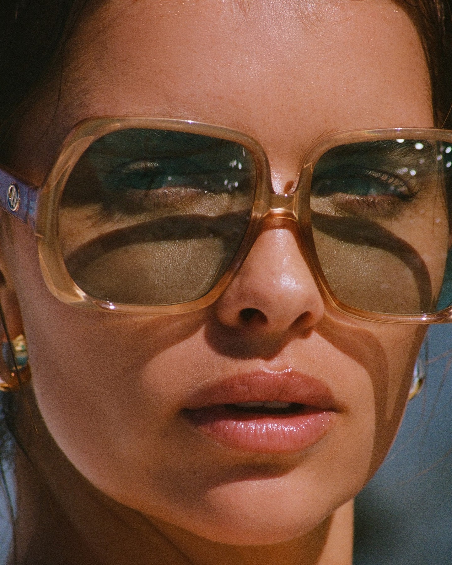 Close-up of a woman wearing oversized sunglasses with a nude lip and gold hoop earrings. The image is sun-drenched and warm.