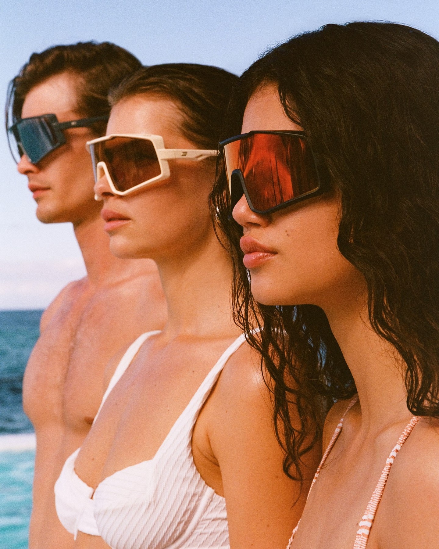 Three people wearing fashionable sunglasses pose outdoors near the ocean on a sunny day; two women wear bikinis, one man is shirtless.